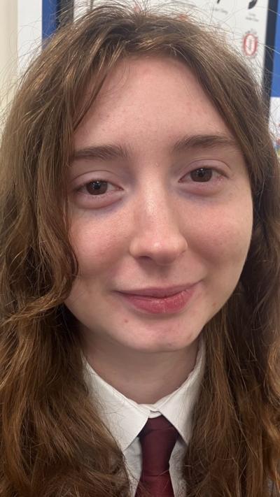 Polly is a young student. She is standing in front of a blue classroom board display and is smiling at the camera. She has light coloured, long hair.