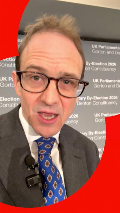 Chris Mason, wearing a suit with a blue patterned tie, speaking into his phone selfie-style with a grey wall in the background.