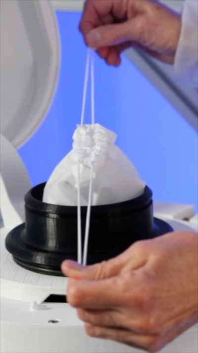 Two hands are pulling tight on the drawstrings of a waste bag. It is in the cylindrical opening of a waste disposal machine