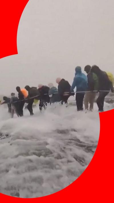 Climbers on a mountain side hold onto a rope while flood water rushes past
