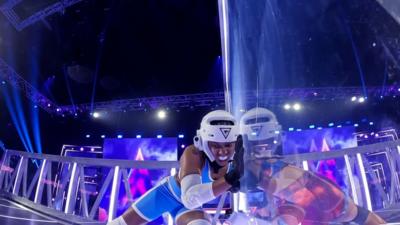 Emily Bell taking part in a challenge on Gladiators. She is wearing a blue outfit and wearing a white helmet and gum shield.