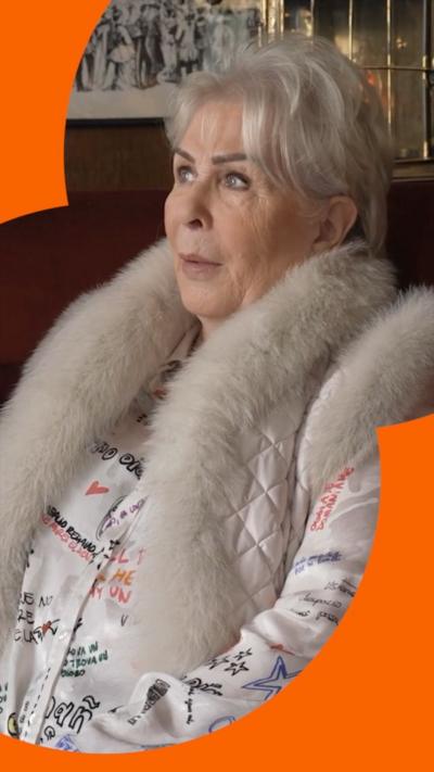Woman with white hair and a jacket with a white fur lined collar looks off to her right