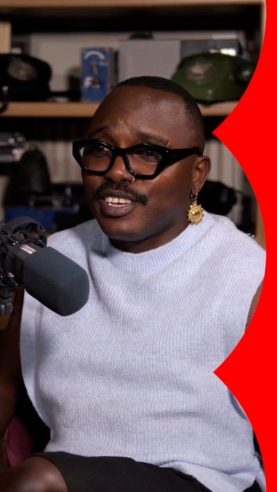 A man in a white vest top sits in front of a microphone with a bookcase behind him