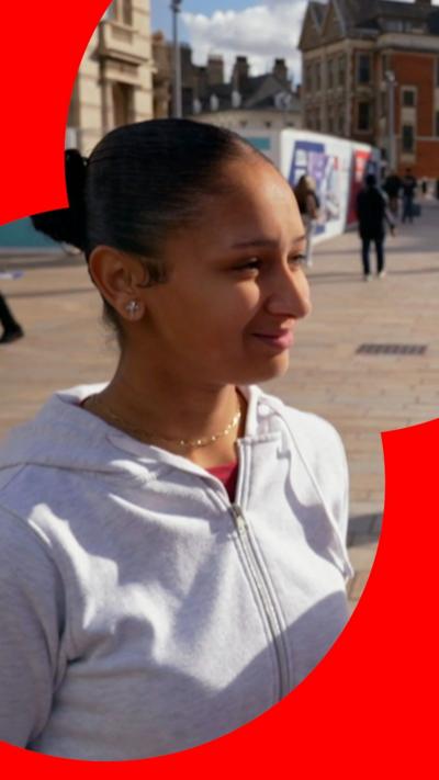 A woman smiles standing in a street in Hull. She has black hair tied in a bun and wears a grey hoodie 