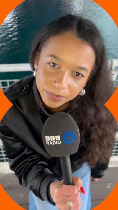 A woman with long dark curly hair holds a Radio 4 microphone. She is wearing blue trousers and a black jacket.