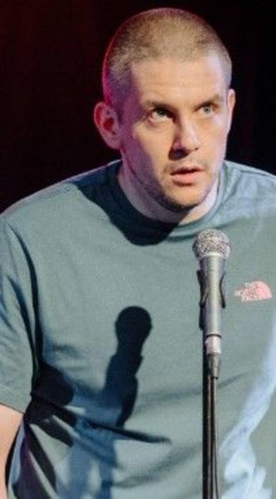 A closeup of a man with a buzz cut standing in front of a microphone, wearing a pale green North Face t-shirt. He looks out at the crowd, backed by total darkness. He looks apprehensive, likely about to drop the punchline of a joke.