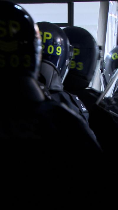 Police officers wearing black tactical clothing and helmets with yellow writing, carry riot shields as they enter a house