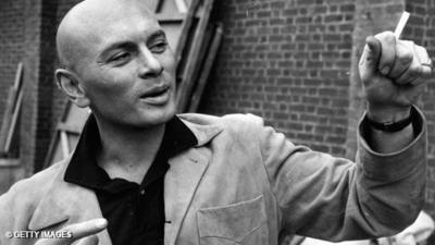 Black and white image of Yul Brynner. He is dressed in a black shirt with a suede looking jacket and is gesticulating like he is in conversation with someone or telling a story. His right hand is pointing like he is miming movement, and points towards his left hand which has a clinched fist with a cigarette sticking out from it. He has a slight smirk on his face.