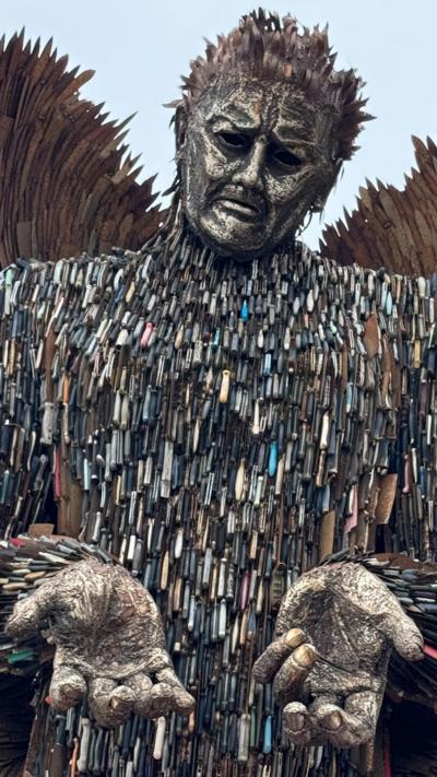 A statue of an angel with its hands out. It is made of knives and metal. There is a blue sky in the background. There is a BBC logo in the top left. 