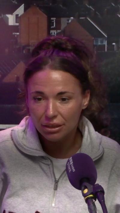 A woman in a grey top in a radio studio sat behind a purple microphone. She has brown, tied-back hair