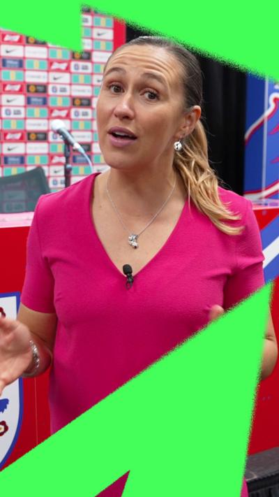 Sarah Dawkins, wearing a pink dress, standing in front of the table with microphones and sponsor logos in the England press room