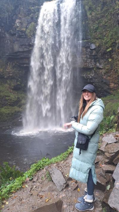 A woman in a long coat and wearing a baseball cap stands on ground next to a large waterfall.