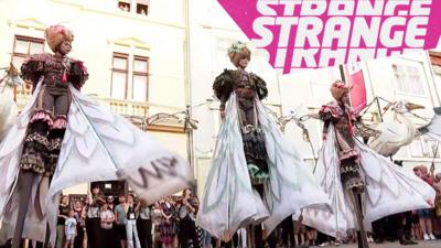 People in bird costumes on stilts and the Strange News logo