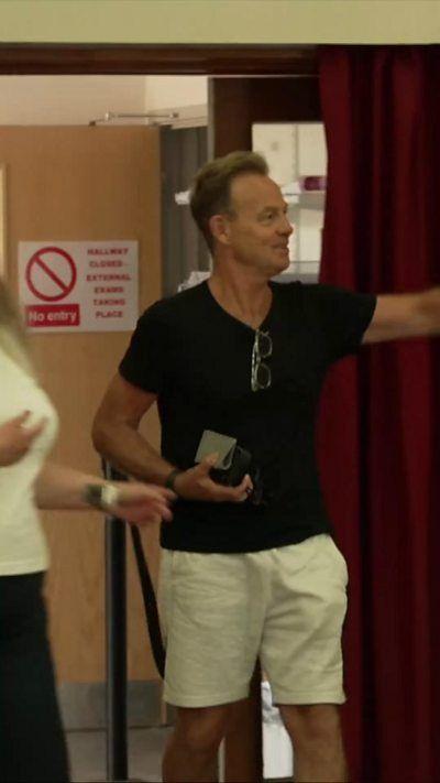 A man with a black t-shirt and shorts on pointing to the side of the frame.
