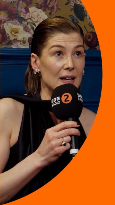 Rosamund Pike wears a black sleeveless dress, holding a Radio 2 microphone.