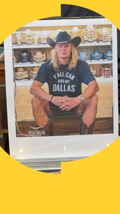 Man in cowboy hat and 'Y'All Can Kiss My Dallas' shirt in front of shelves of cowboy hats