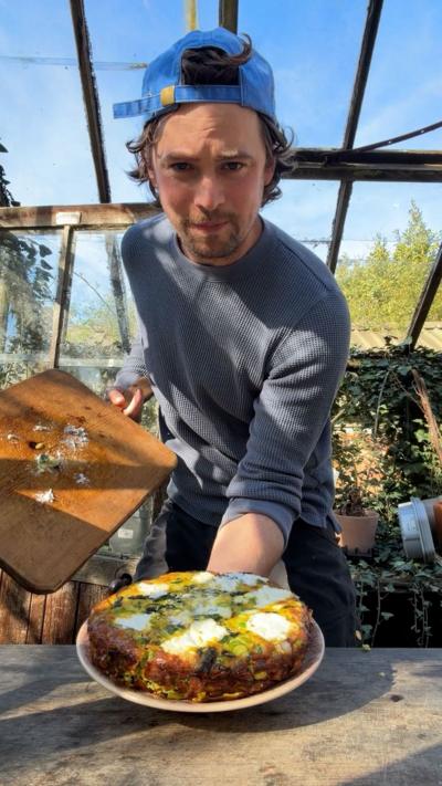 Julius Roberts presents a freshly cooked frittata on a plate. He is wearing a blue grey top and backwards blue baseball cap.