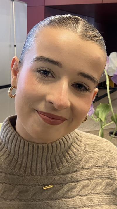 A young woman with dark hair, tied back, looks into the camera. She is wearing a beige jumper and gold, hoop earrings.