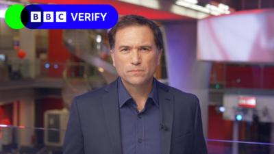 Ros Atkins in a dark jacket and dark shirt looks directly at the camera, with a number of screens and the red walls of the BBC newsroom visible behind him.