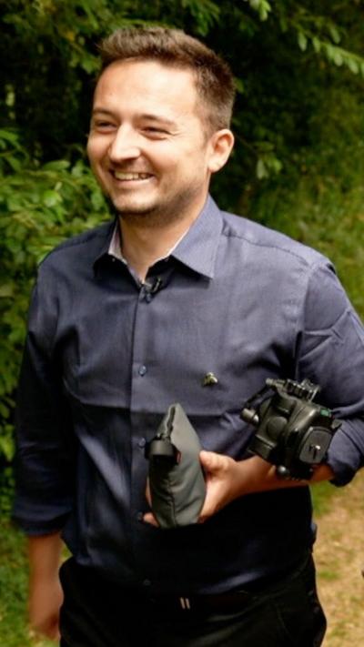A man is wearing a blue shirt and is holding a camera while walking in wildlife.