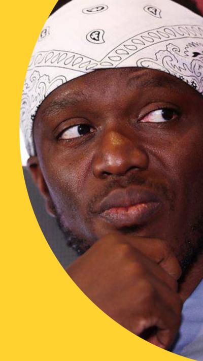 KSI looks to the side with his hand on his chin. He wears a white and black bandana 