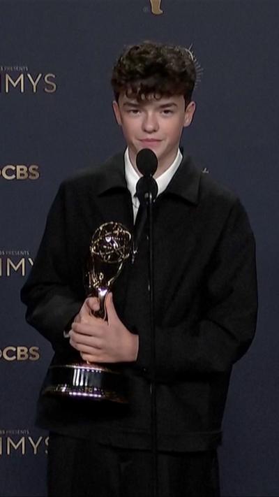 Owen Cooper, wearing a dark suit and tie, holds an Emmy Award in front of a microphone.