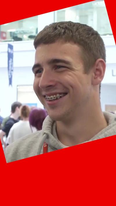 A student smiles - he has short, light brown hair and wears a grey hoodie