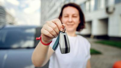 A woman in a white t shirt with a bob holds car keys to the camera, with a car behind her.