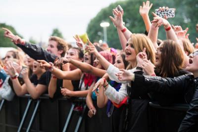 A crowd of people hanging over a barrier at a concert with their hands in the air, singing along. The crowd is mainly made up of young people with different coloured clothing on and different hairstyles.