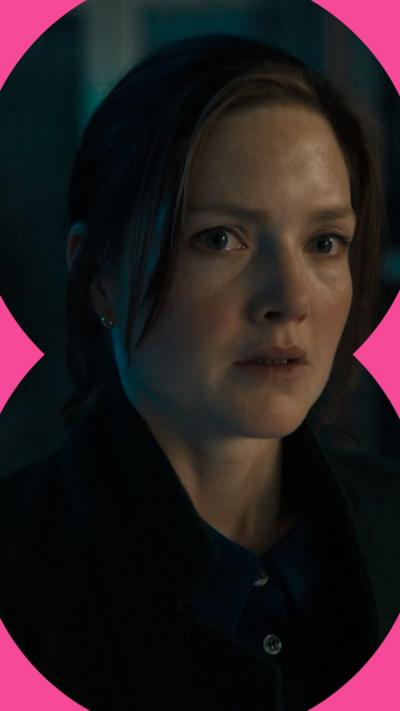 A woman in a dark interior space is wearing a black jacket and looks scared.