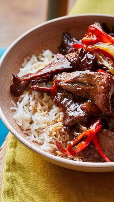 A bowl of cooked beef and peppers in a dark sauce with rice alongside a bowl of chopped cucumber sprinkled with sesame seeds