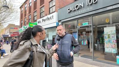 A woman wearing a leather jacket interviews a man in a padded vest outside a hairdressing shop in a busy urban street.