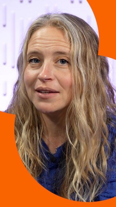 A woman with long blond wavy hair wearing a blue top, looks at the camera