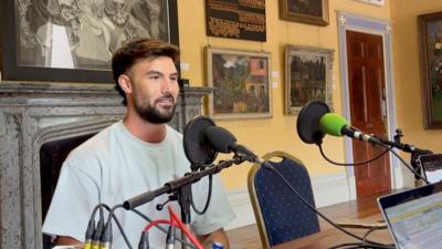 A man with short brown hair and a beard, wearing a light blue t shirt speaks in front microhphones