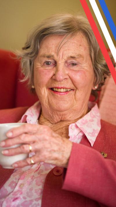 Prudence sits on a chair in her living room with a cup of tea. She is wearing a pink suit and pink lipstick.