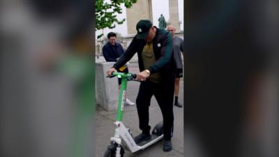 A man has one foot on a green and white electric scooter and one foot on the pavement. He wears a baseball cap 