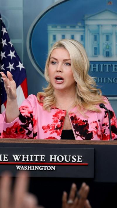 Karoline Leavitt stands behind a podium wearing a bright green jacket as she speaks to press. 