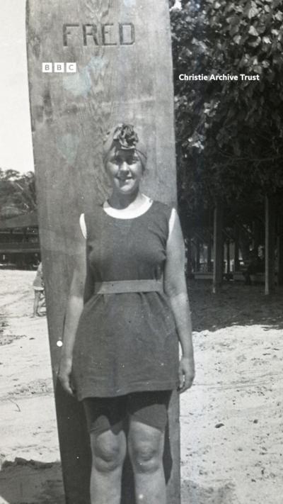 A young Agatha Christie stands on a beach against a large surf board.