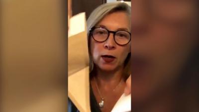 A woman with black glasses and grey hair holds up a triangular piece of material to camera