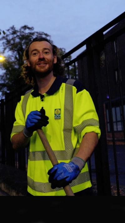 Eddie, a Glasgow street cleaner, who cleans the city's streets during the night. He is smiling at the camera and is wearing a yellow high vis jacket.