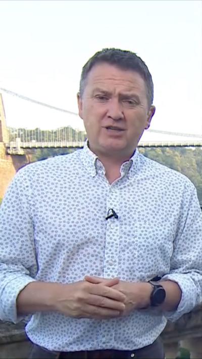 BBC Weather presenter Matt Taylor wearing white and blue shirt with small flowers. He is standing in front of Clifton Bridge