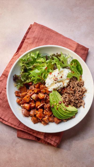 A white plate sitting on a napkin with hot honey beef, sweet potato, avocado, cottage cheese and spinach on it