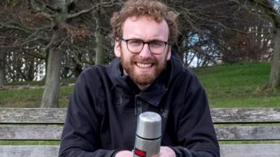 Will Sheers, a bearded man with glasses, sat on a park bench and holding a flask of hot tea.