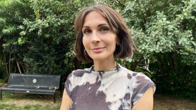 Milly stands in the gardens of the Trinity Centre. She has short dark-brown hair and wears black and white short-sleeved top.
