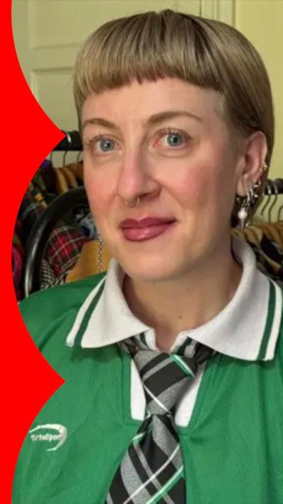 A blonde woman wears a green and white collared shirt with a checked tie. Behind her are racks of clothes 