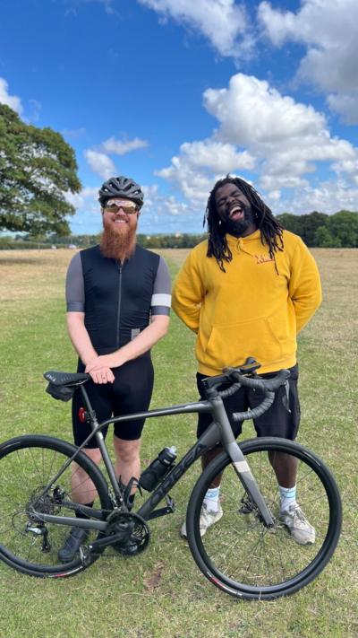 Two men standing in front of a bicycle, in a field with a large tree in the distance and a bright blue sky with fluffy white clouds. The man on the left is wearing cycling gear and the man on the right is wearing a yellow top and black shorts.