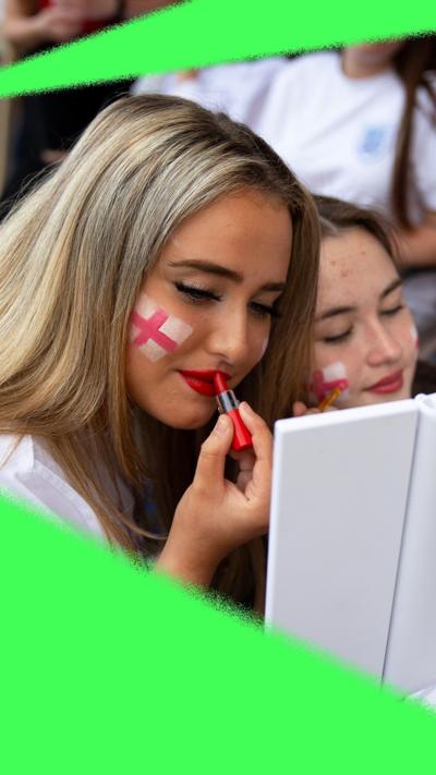 A girl with an England flag painted on her face, applying red lipstick with her friend in front of a mirror during a football watch party