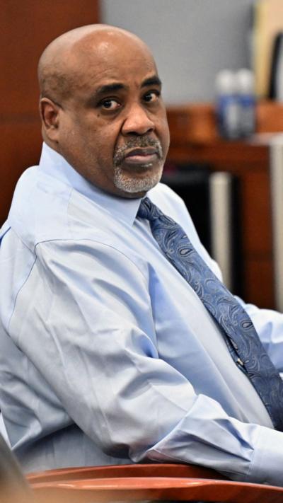 A man wears a light blue button up and blue paisley tie as he sits turned facing the camera inside of a courtroom.