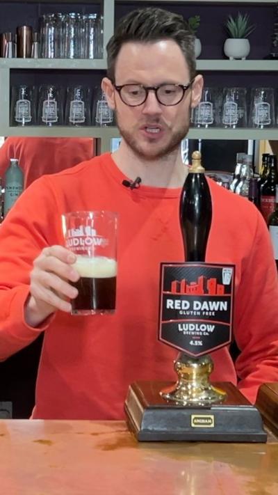 A man with a beard and glasses stood behind a bar of a pub with a half pint of beer. He is wearing an orange jumper