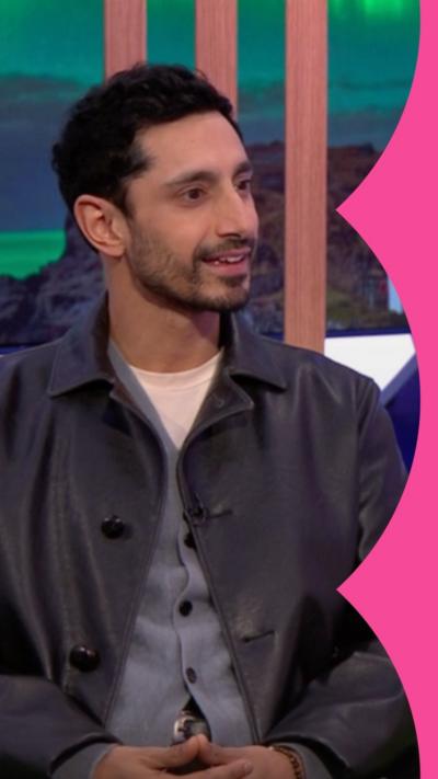 A man, Riz Ahmed, with short black hair, sits on a sofa. He wears a black leather jacket